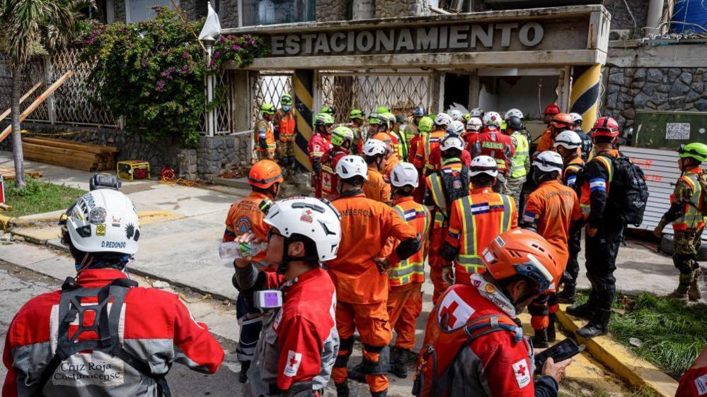 Rescue workers from Mexico, El Salvador, and Costa Rica are standing in front of the entrance to the site of a collapsed multi‑storey car park