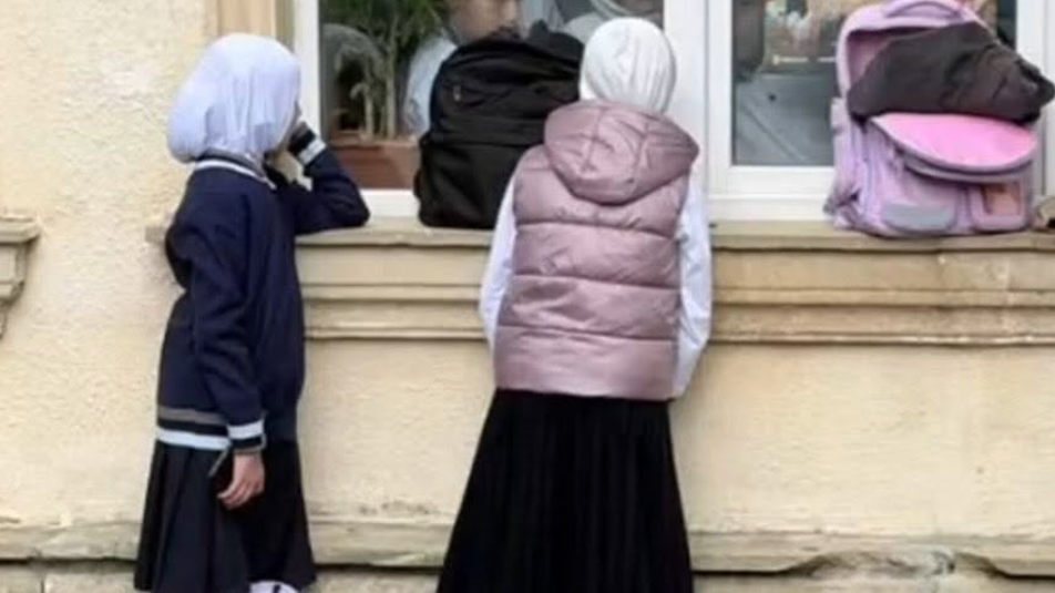 Two hijab‑wearing schoolgirls are peering through a school window, with a pink schoolbag placed on the window ledge next to them.