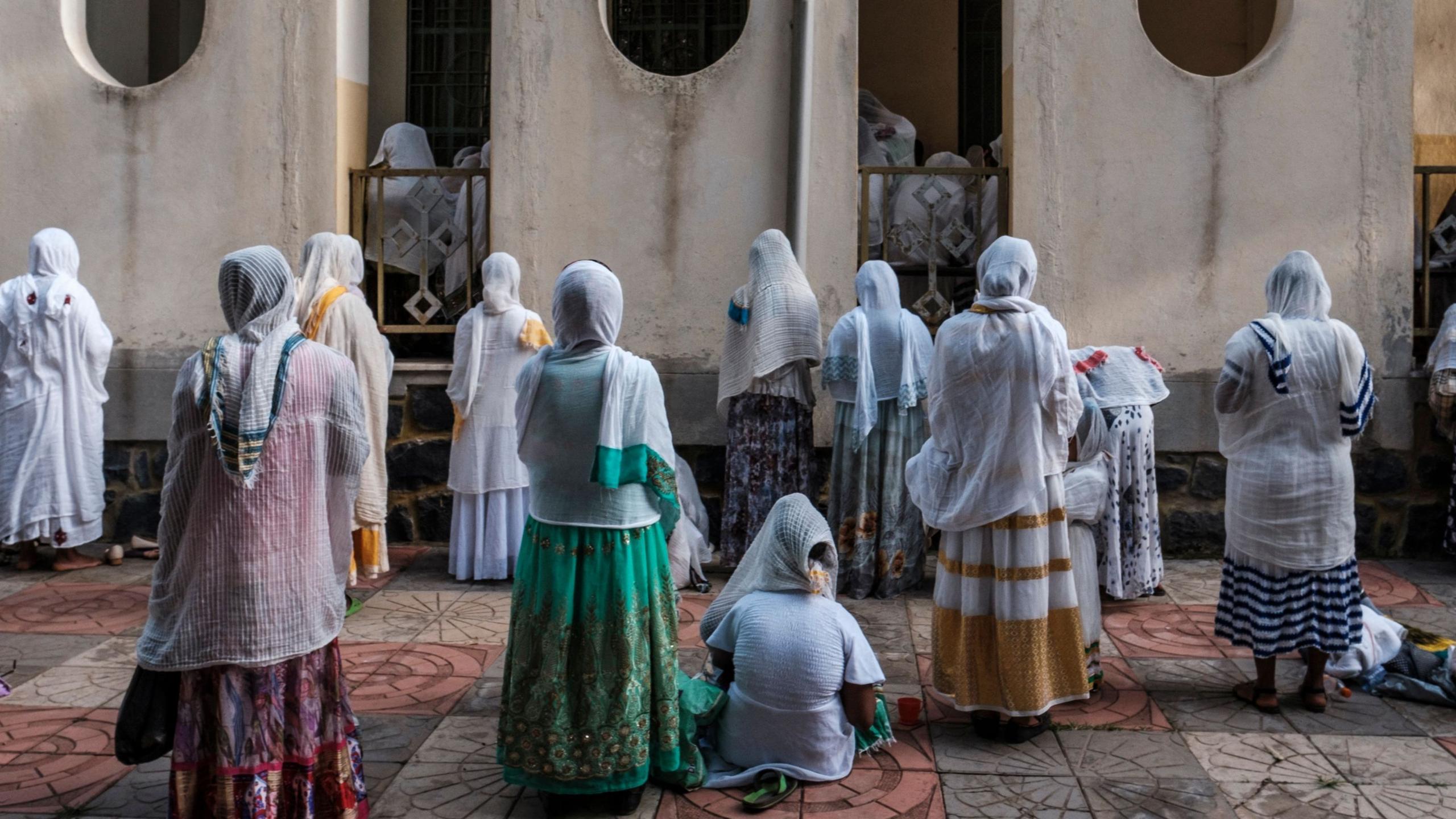 Women wearing colourful traditional Ethiopian dresses pray outside a church. Some stand while others kneel.